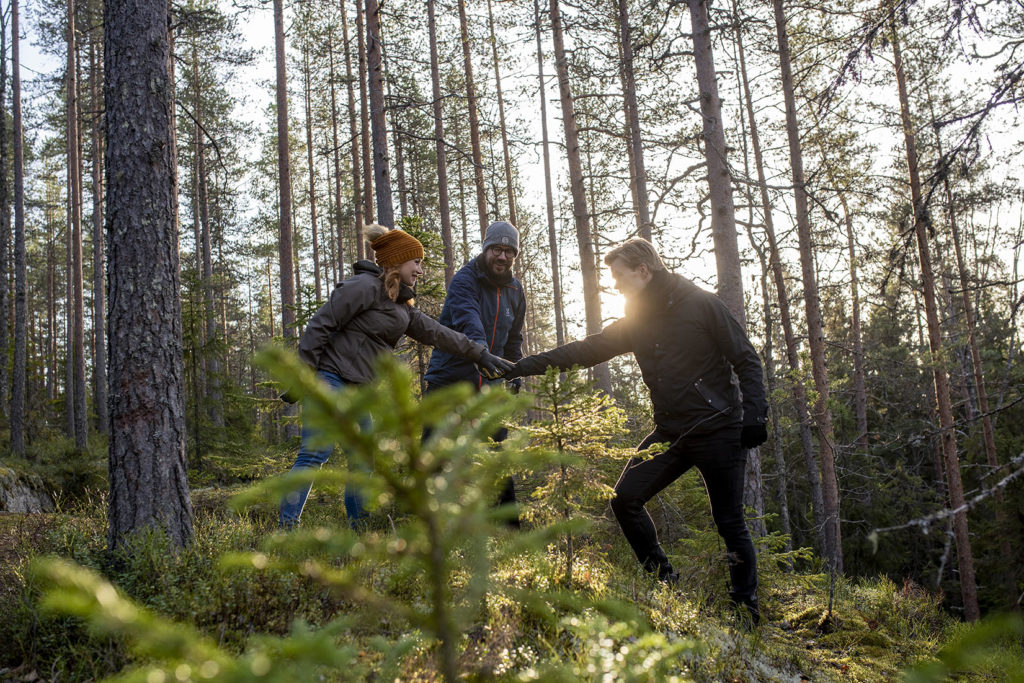 Kolme hymyilevää ihmistä syksyisessä metsässä. Jokainen on ojentanut käden päällekkäin ringin keskelle. Aurinko paistaa puiden lomasta.