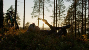 Mies makaa metsän mättäällä, metsän reunalta häikäisevä aurinko luo siluetin henkilölle.