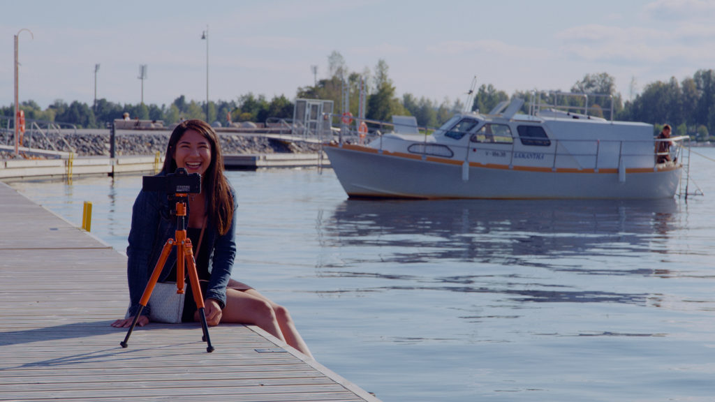 Nainen istuu kameran kanssa sataman kivireunuksella, taustalla kimmeltävää vettä ja veneitä